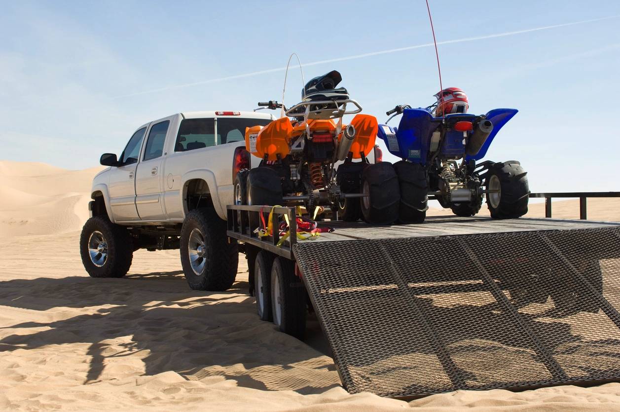 A large truck towing a trailer with two colorful ATVs parked on sand dunes.